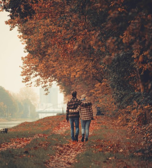 A couple walking arm in arm along a riverside path lined with autumn trees