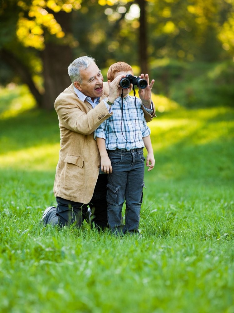 A grandfather kneeling in the grass beside his grandson, helping him look through a pair of binoculars