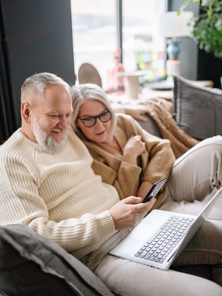 An older couple sitting together on a sofa, looking at a laptop and a phone